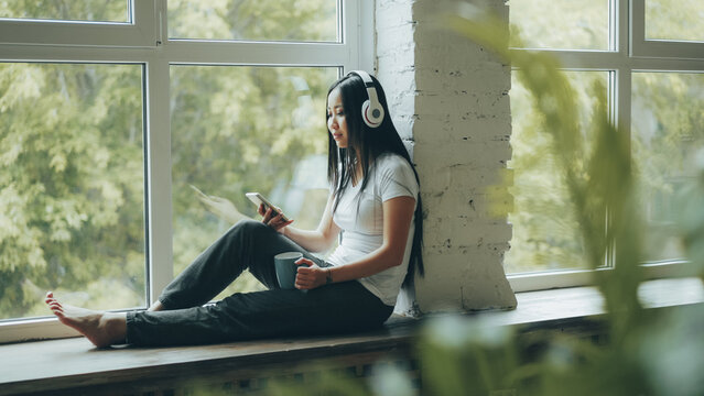 Pretty Asian student is enjoying music in headphones sitting on window sill and using smartphone touching screen. Modern youth lifestyle and technology concept.