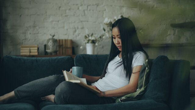 Clever young lady with long dark hair is reading book and holding cup of coffee sitting on couch in living room in beautiful flat enjoying solitude and peace at weekend.