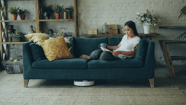 Beautiful Asian girl is reading book resting on sofa in living room while robotic vacuum cleaner is dry cleaning floor and carpet. Technology and young people concept.
