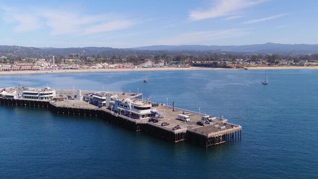 Aerial view flies over the wooden pier of Santa Cruz wharf, revealing the sandy beach, amusement park boardwalk, calm Pacific ocean waters, and distant hills on a sunny day in California.