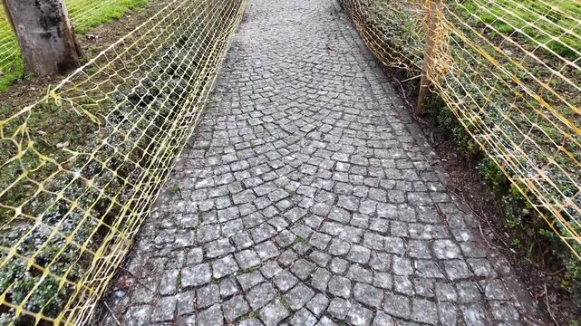 Point of view walking shot along narrow cobblestone path with safety netting fences in city park garden.