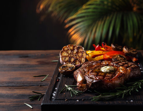 Grilled steak sits on dark wooden board. Roasted garlic and colorful peppers add rustic charm. Fresh rosemary sprigs frame the dish with earthy scent. Warm, golden light highlights savory textures