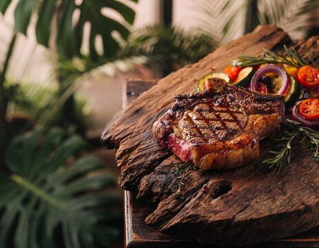 Grilled steak rests on rustic wooden board. Fresh herbs and vibrant veggies surround the meat. Warm lighting enhances rich, savory tones. Tropical leaves frame the scene in soft green