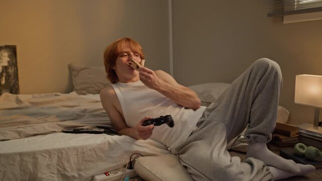Caucasian young man lying on floor, eating takeaway pizza and playing video games with gamepad. Perfect for youth lifestyle commercials, fast food delivery promotions, gaming advertising