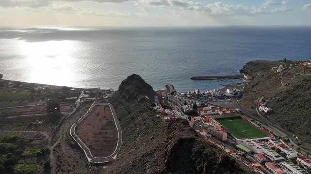 Aerial drone push in shot revealing Roque de la Cruz mountain, the vibrant coastal town Playa de Santiago, and harbor on La Gomera. Beautiful sunrise light illuminates the Atlantic Ocean.