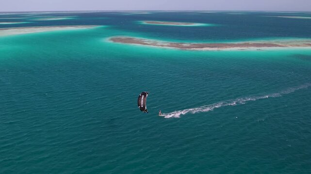 Aerial view of a kitesurfer gliding across the turquoise waters of the Los Roques archipelago with coral reefs visible below in Los Roques, Dependencias Federales, Venezuela.