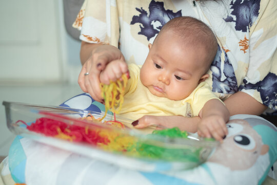 Asian Baby Grabbing Colorful Spaghetti Noodles in Mother Lap During Tactile Sensory Game