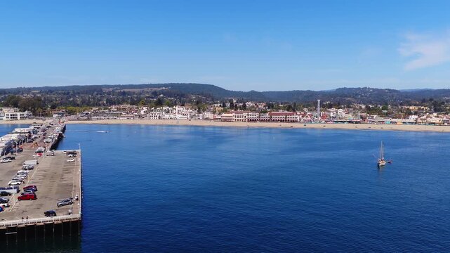 Aerial panorama of the Santa Cruz coastline in California showing the sandy beach, Beach Boardwalk amusement park, wharf pier, and a sailboat anchored in Monterey Bay on a clear day.