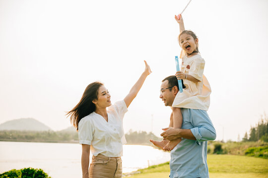 Asian family child girl piggyback riding back on father raising hands like flying outdoor nature park with mother, Happy parent daughter having fun together