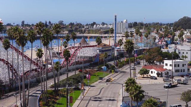 Aerial view tracks along the Giant Dipper roller coaster, sandy coast, palm trees, and ocean amusement park attractions in Santa Cruz, California, on a sunny day.