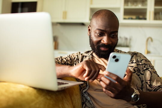 Bearded man texting on smartphone beside laptop at home