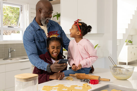 African American family making christmas cookies in kitchen, dad holding smartphone, kids laughing