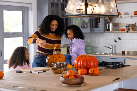 African American mother and two child girls carving pumpkins at kitchen island for halloween