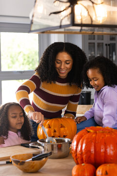 African American mom with youth daughters carving pumpkin and scooping pot in kitchen for halloween