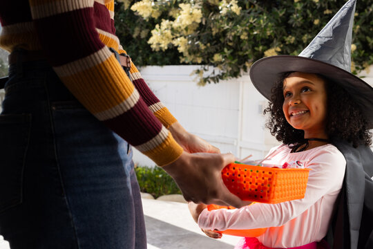 African female adult handing orange pail to youth in witch hat holding pumpkin front yard halloween