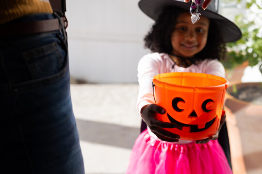 African woman handing halloween candy on porch, child holding orange bucket wearing witch hat, tutu