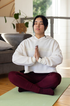African American woman meditating on pale green yoga mat at home, in white hoodie