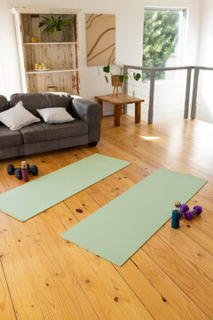 Two mint-green yoga mats are lying parallel on varnished wooden floor near dumbbells, water bottles