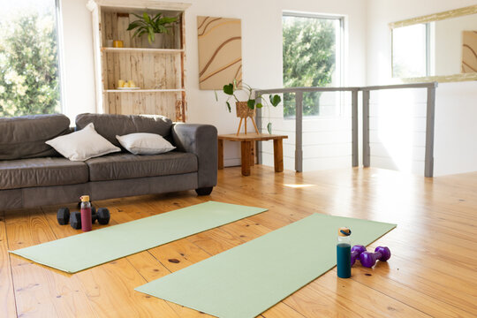 Two light-green yoga mats are lying on hardwood floor in sunlit loft with dumbbells and bottles