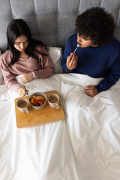 Diverse couple wearing mauve top and navy sweatshirt sitting in bed sharing fruit from wooden tray