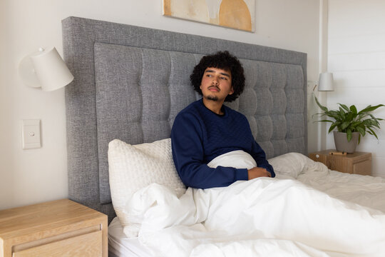 African American man sitting against gray tufted headboard on bed at home, wearing navy sweatshirt