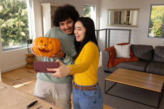 Diverse couple posing with carved pumpkin and smartphone in living room halloween