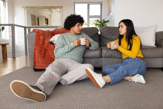 Diverse couple sitting on gray carpet in front of sectional sofa, holding ceramic mugs and talking