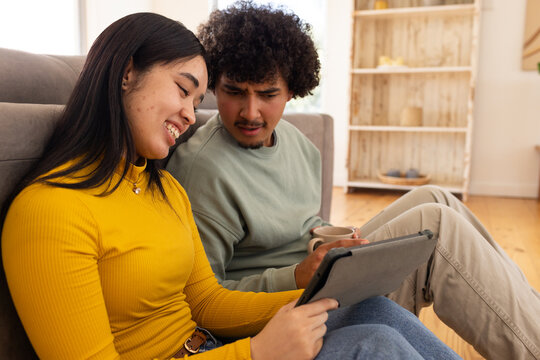 Diverse couple holding tablet on gray couch in living room, mustard top and sage-green sweatshirt