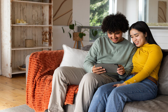 African American couple sitting on gray upholstered couch at home, holding tablet device