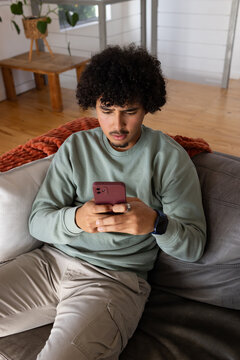 African American man sitting on gray sofa at home, holding maroon smartphone, wearing smartwatch