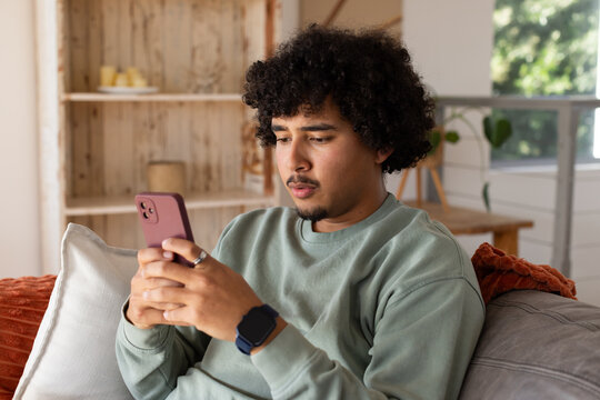 Adult African man sitting on couch at home wearing pale-green sweatshirt holding pink smartphone