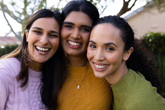 Diverse female friends leaning close and smiling in garden wearing knit sweaters and pendant