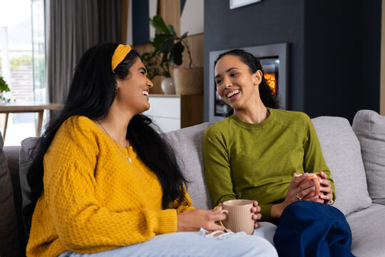 African American and Indian women friends sitting on sofa at home wearing sweaters holding mugs