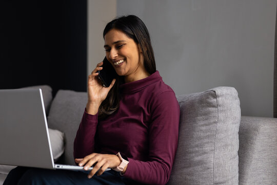 Woman calling on phone while typing on laptop on sofa wearing maroon turtleneck and pale smartwatch