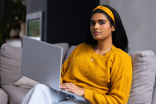 Woman sitting on grey sofa at home working on silver laptop wearing mustard sweater and headband