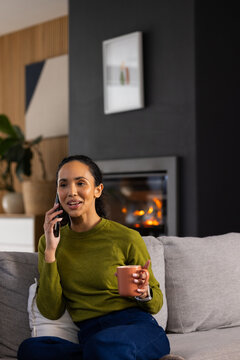 African American woman sitting on sofa in green sweater holding terracotta mug and talking on phone