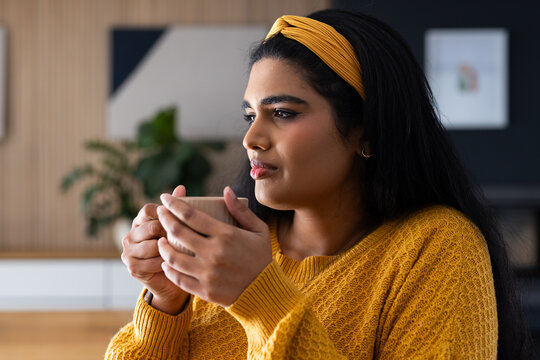 Indian woman wearing mustard sweater, yellow headband, holding ceramic mug, gazing right in kitchen