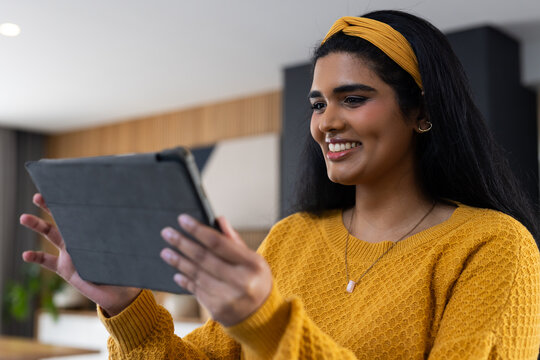 Indian female holding tablet in folio case, wearing mustard sweater and headband, looking at screen
