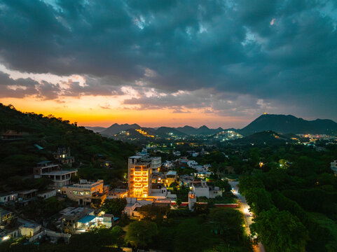 Aerial view of illuminated buildings nestled among lush greenery and rolling hills under a dramatic sunset sky in Udaipur, Rajasthan, India.