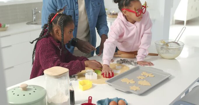 Christmas urging African American man guiding two youth girls cutting dough with cutters at island