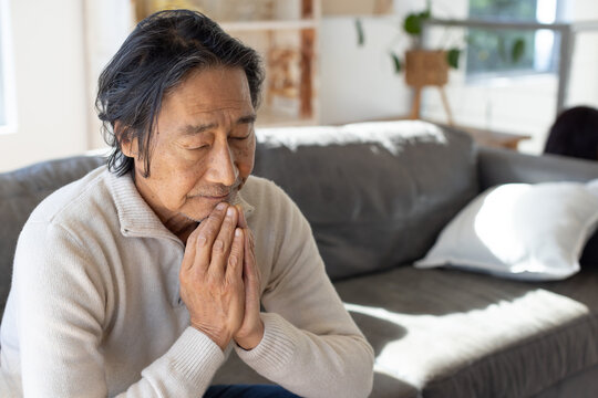 Senior Asian man wearing beige pullover, sitting in living room on gray sofa, clasping chin
