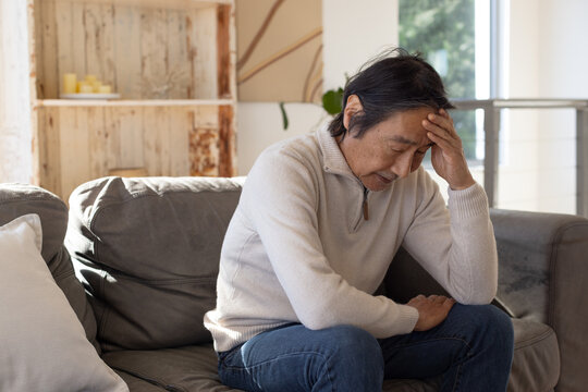 Senior Japanese man holding forehead on grey couch at home, in beige sweater and jeans