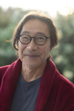 Senior Asian man wearing round black eyeglasses, red robe jacket and gray shirt, smiling in garden