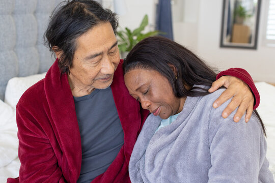 Senior Japanese man holding and comforting African woman wearing plush robes while sitting on bed