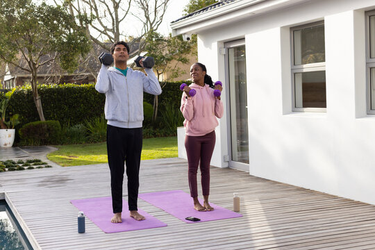 African American couple in hoodies exercising on purple mats on deck beside pool with dumbbells
