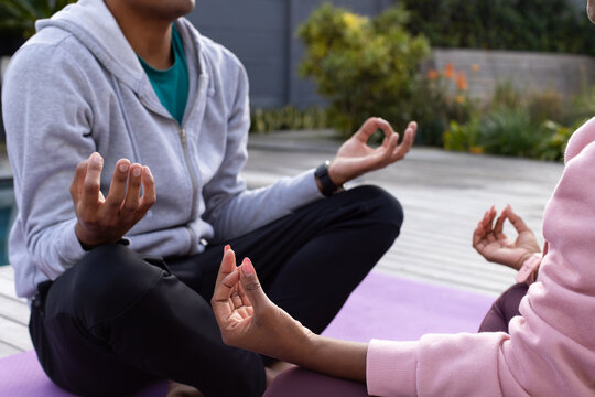 African American couple meditating on mat on deck, wearing gray hoodie, pink sweatshirt, smartwatch