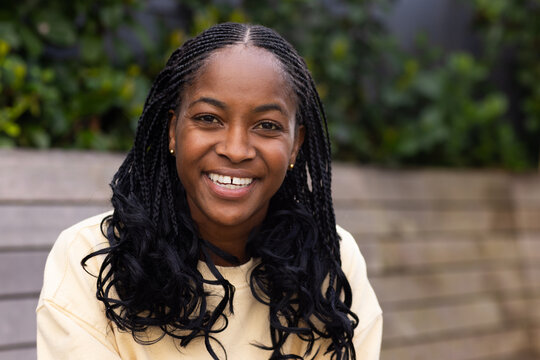 African American woman sitting on wooden slatted bench by planted hedge, wearing cream sweatshirt