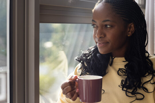 Adult African American woman standing by large window holding mauve mug wearing pale yellow sweater
