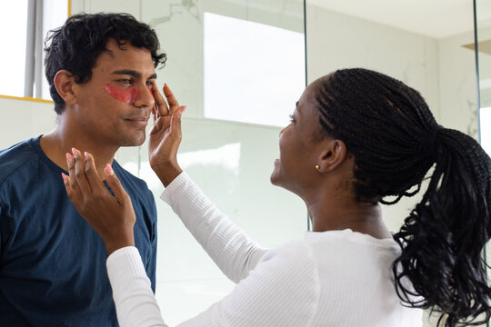 Diverse couple standing in bathroom by mirror, woman applying red under-eye gel patches