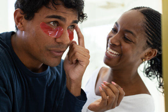 Couple standing at bathroom mirror, man adjusting red under-eye patches, woman in white top smiling